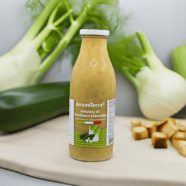 Vellutata bio di zucchina e finocchio AmoreTerra in bottiglia di vetro, ambientata tra zucchine e finocchi freschi su tovaglietta beige, perfetta per comunicare freschezza e naturalità.